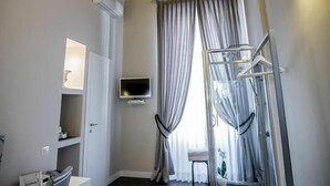 A hotel room with a classic ceiling, a desk with a chair, a TV, a window with curtains, and a small plant.