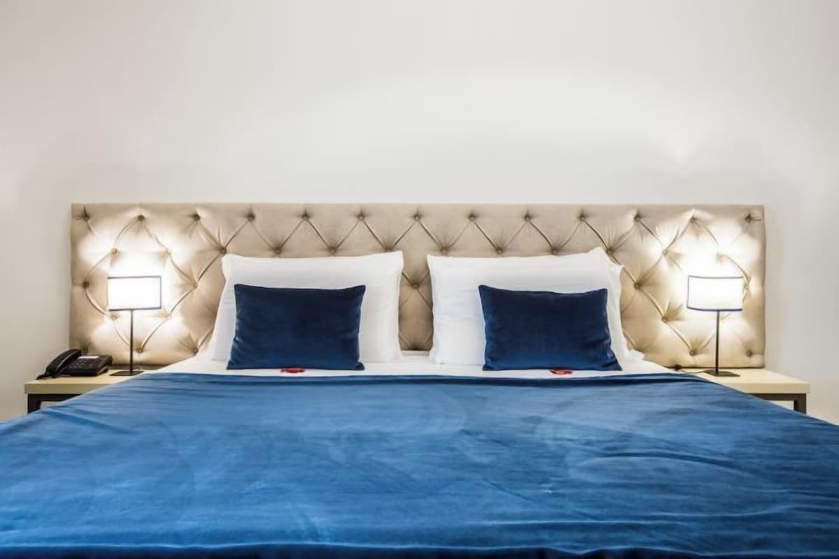 A hotel room with a tufted headboard, two bedside lamps, a blue bedspread, and two pillows.