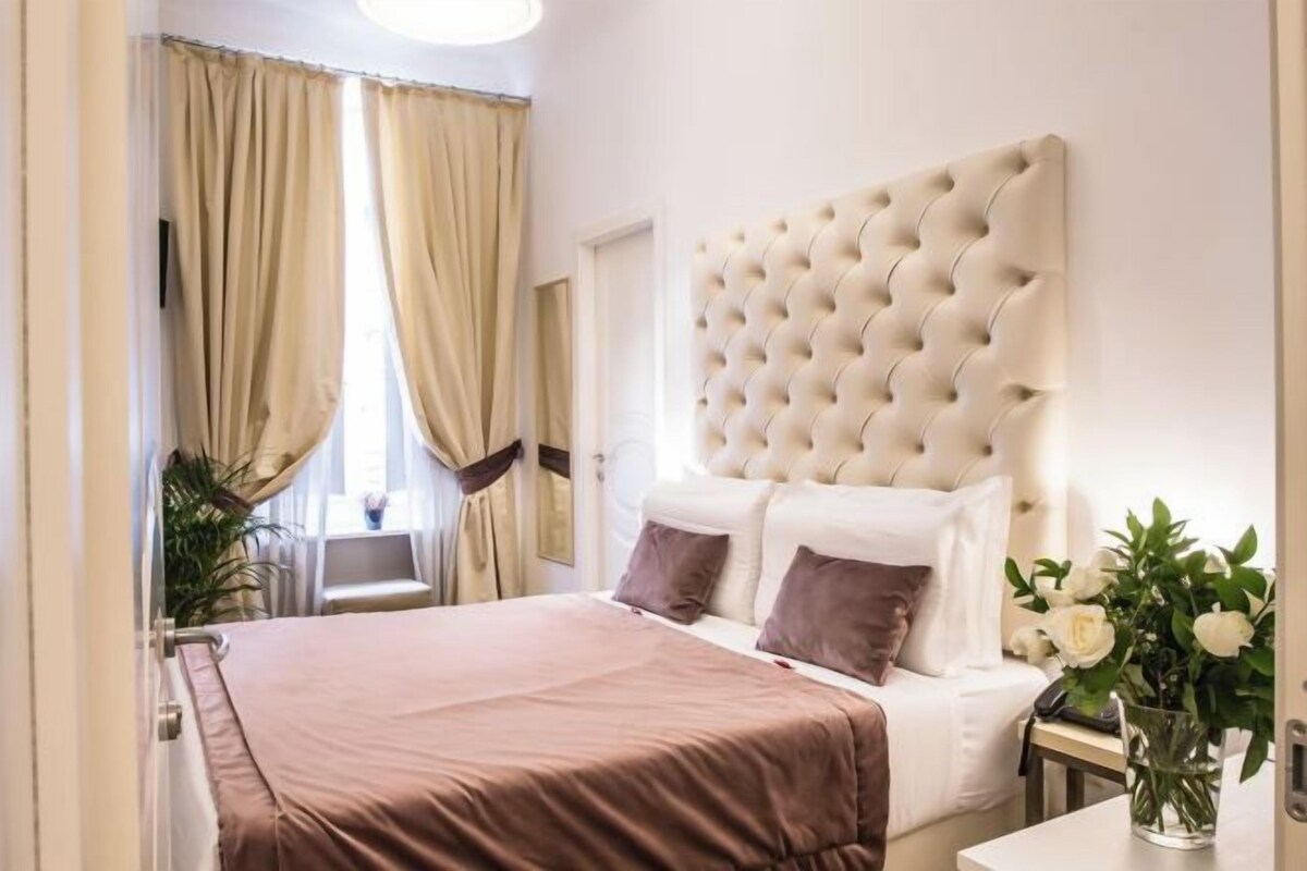 A bedroom with a large bed, a tufted headboard, a bedside table with a vase of flowers, and a window with curtains.