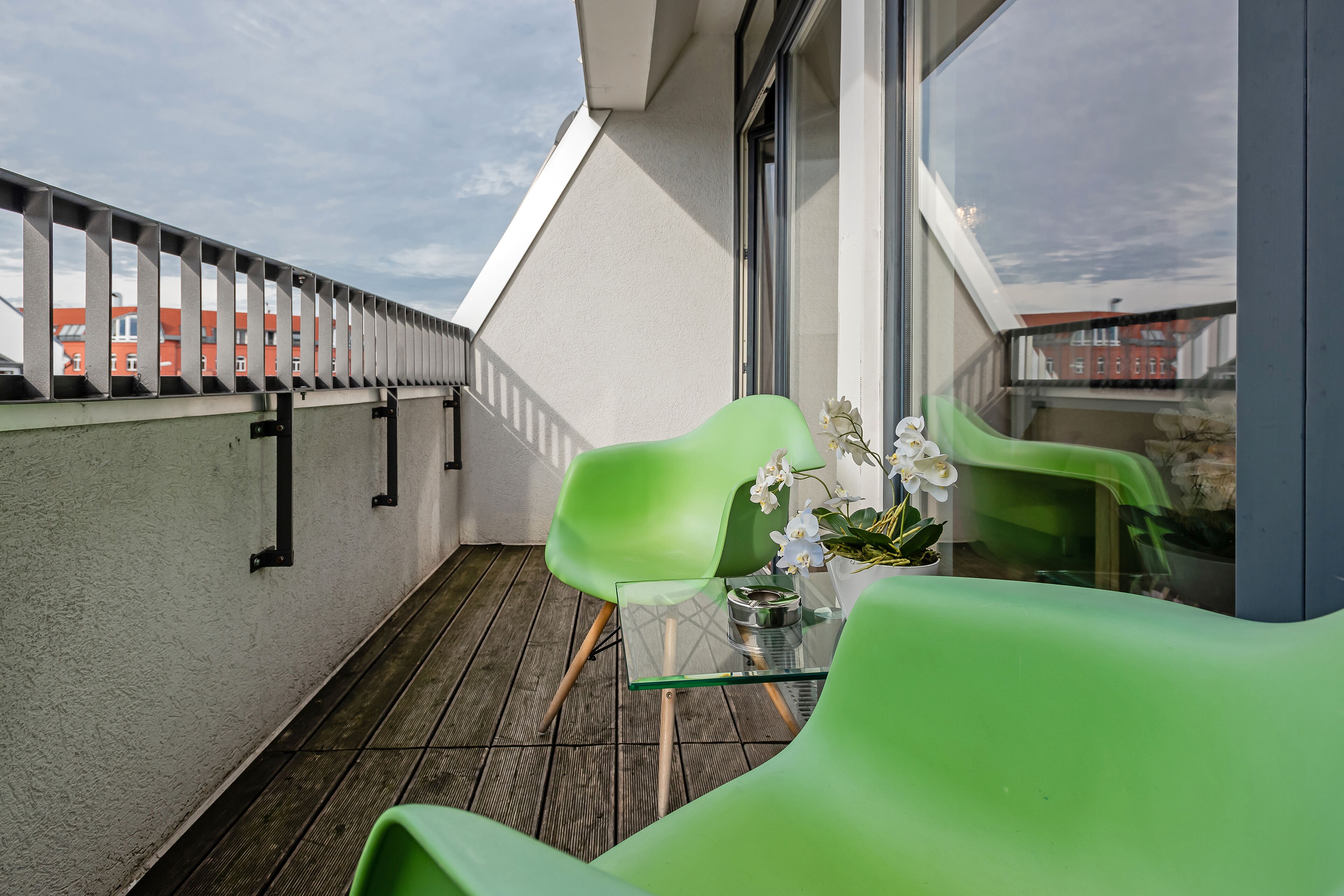 Un balcón con una silla verde, una mesita con una planta y vistas a edificios al fondo.