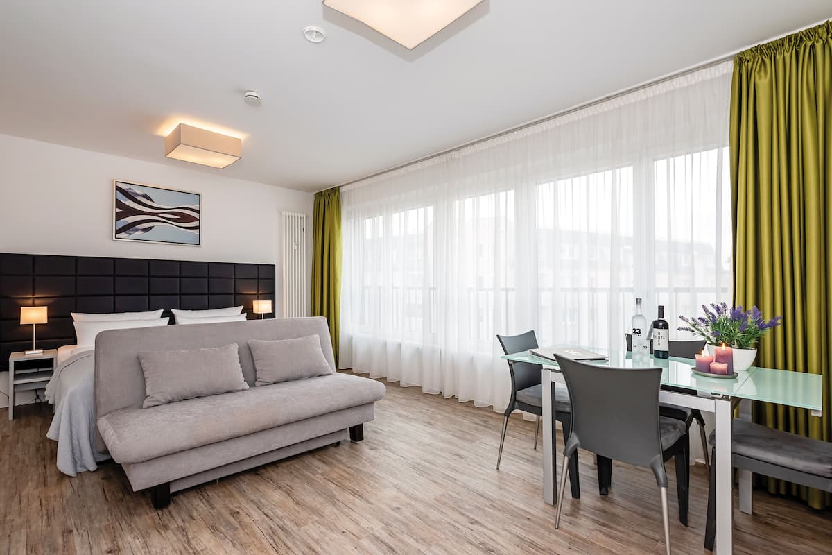 Moderni hotellihuone, jossa on sohva, ruokapöytä tuoleineen, sängynpäätyä koristeleva sänky ja suuri ikkuna, jossa on läpikuultavat verhot.