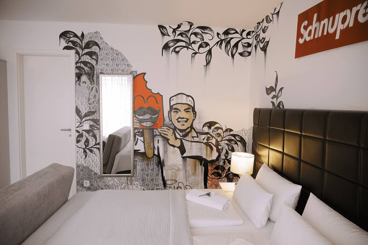 Moderni hotellihuone, jossa on suuri sänky, yöpöytä lampun kanssa sekä seinä, jossa on peili ja taideteos.