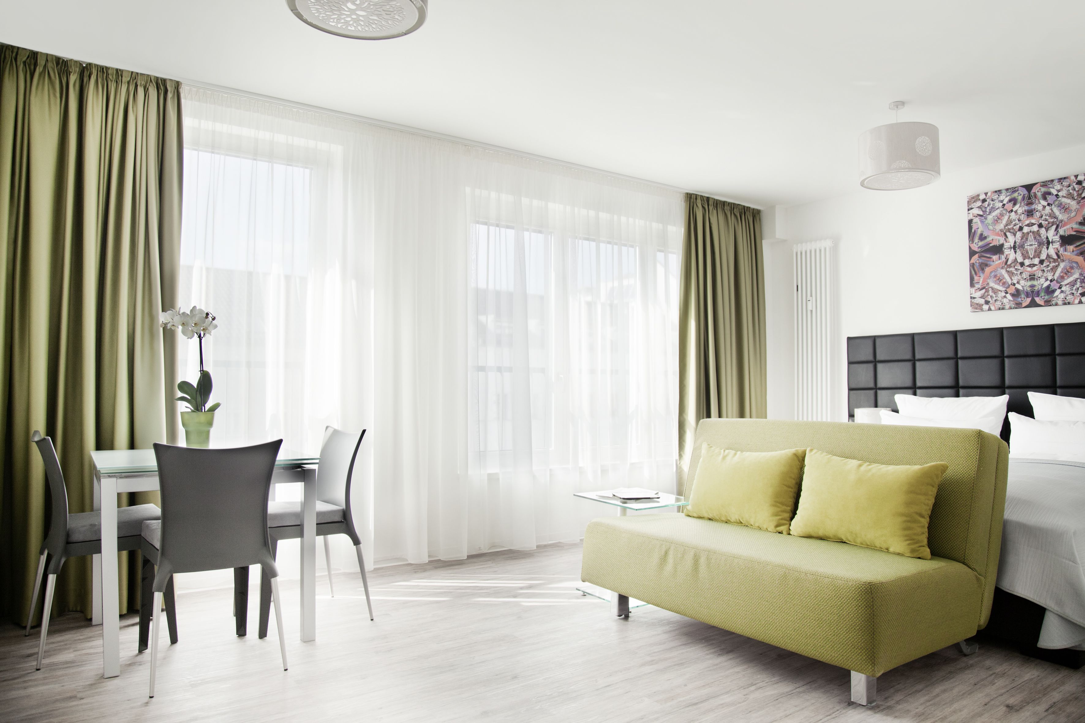 Una habitación de hotel moderna con un sofá verde, una mesa de comedor con sillas y un ventanal con cortinas translúcidas.
