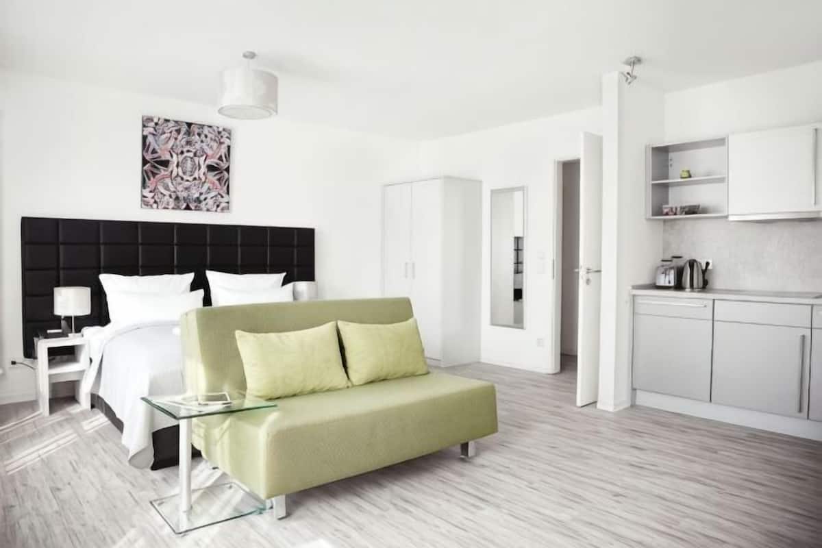 Moderni hotellihuone, jossa on sänky, vihreä sohva, lasinen sohvapöytä, valkoinen yöpöytä, valkoinen lamppu, keittiöalue ja valkoinen vaatekaappi.