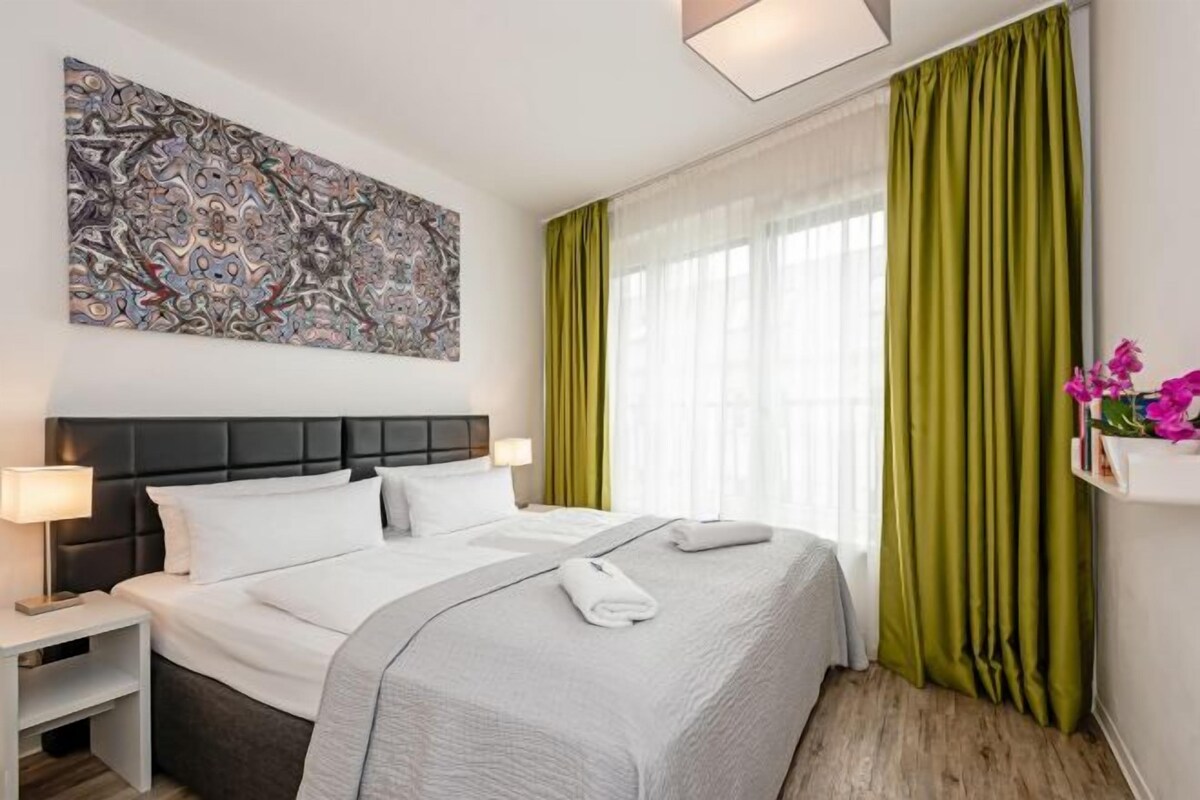 A hotel room with a large bed, a headboard, a nightstand, a wall-mounted abstract painting, a window with green curtains, and a small shelf with flowers.