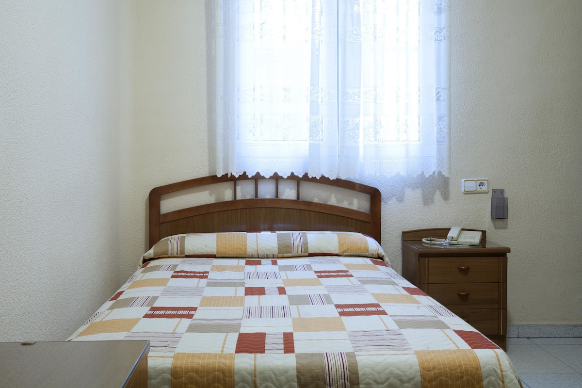Un dormitorio con una cama de madera, un colcha, una mesita de noche con un teléfono y una ventana con cortina.