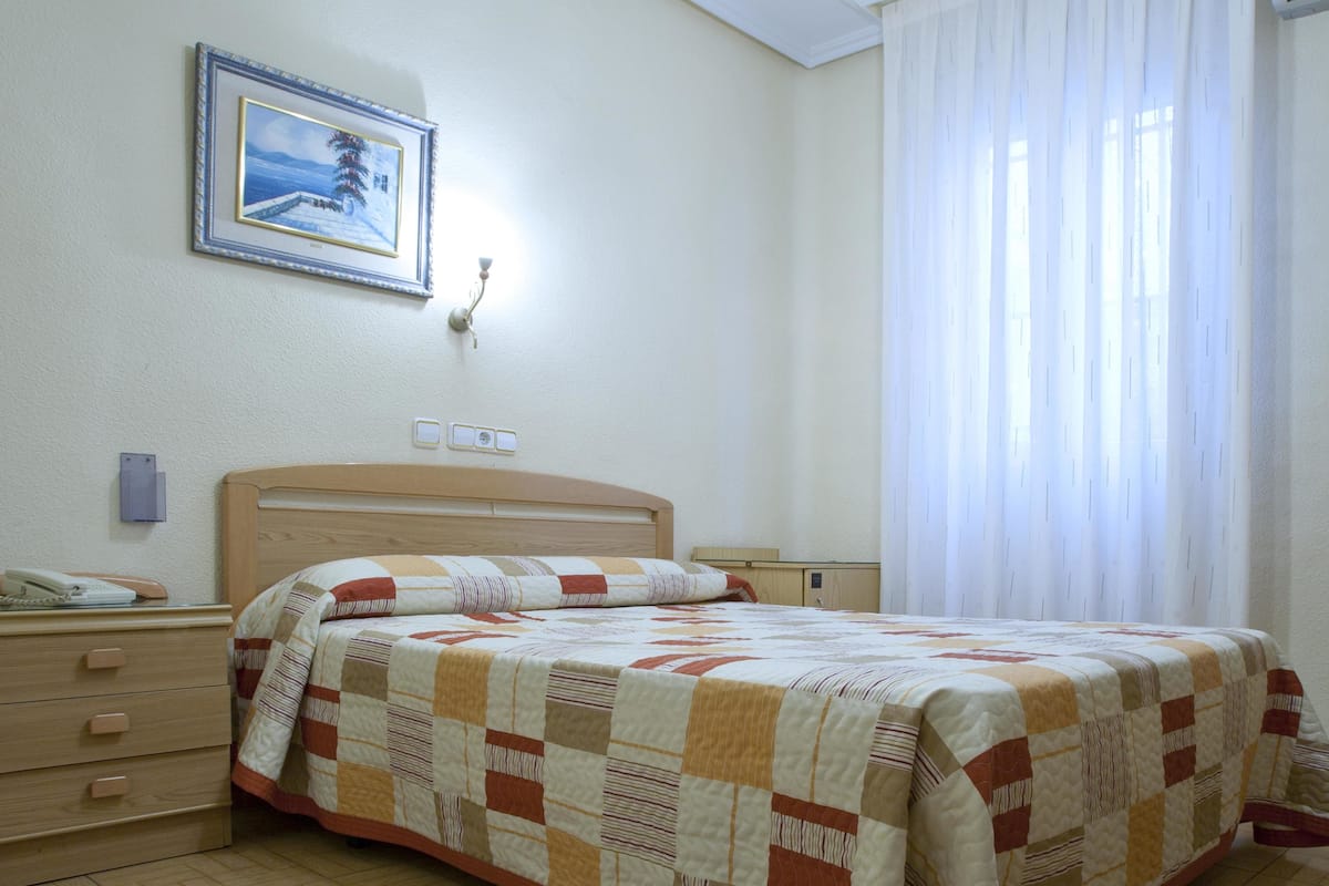 Un dormitorio con una cama de madera, una mesita de noche, un cuadro enmarcado en la pared y una ventana con cortinas translúcidas.