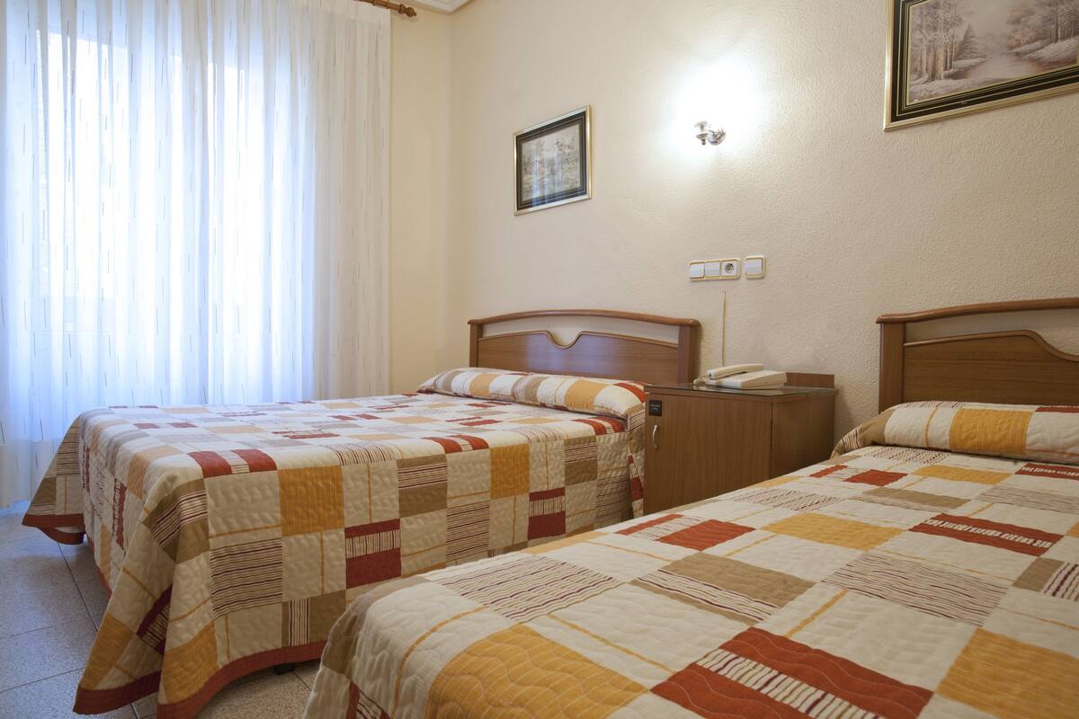 Habitación de hotel con dos camas, cada una con cubrecama acolchado, cabecero de madera y mesita de noche con lámpara.
