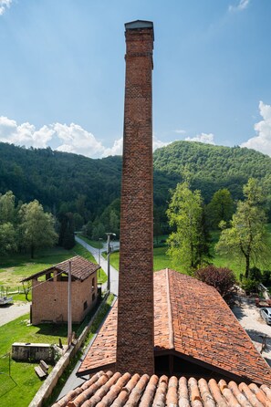 Un alto chimenea de ladrillo, un edificio de ladrillo con tejado de tejas y un paisaje verde con árboles y colinas al fondo.