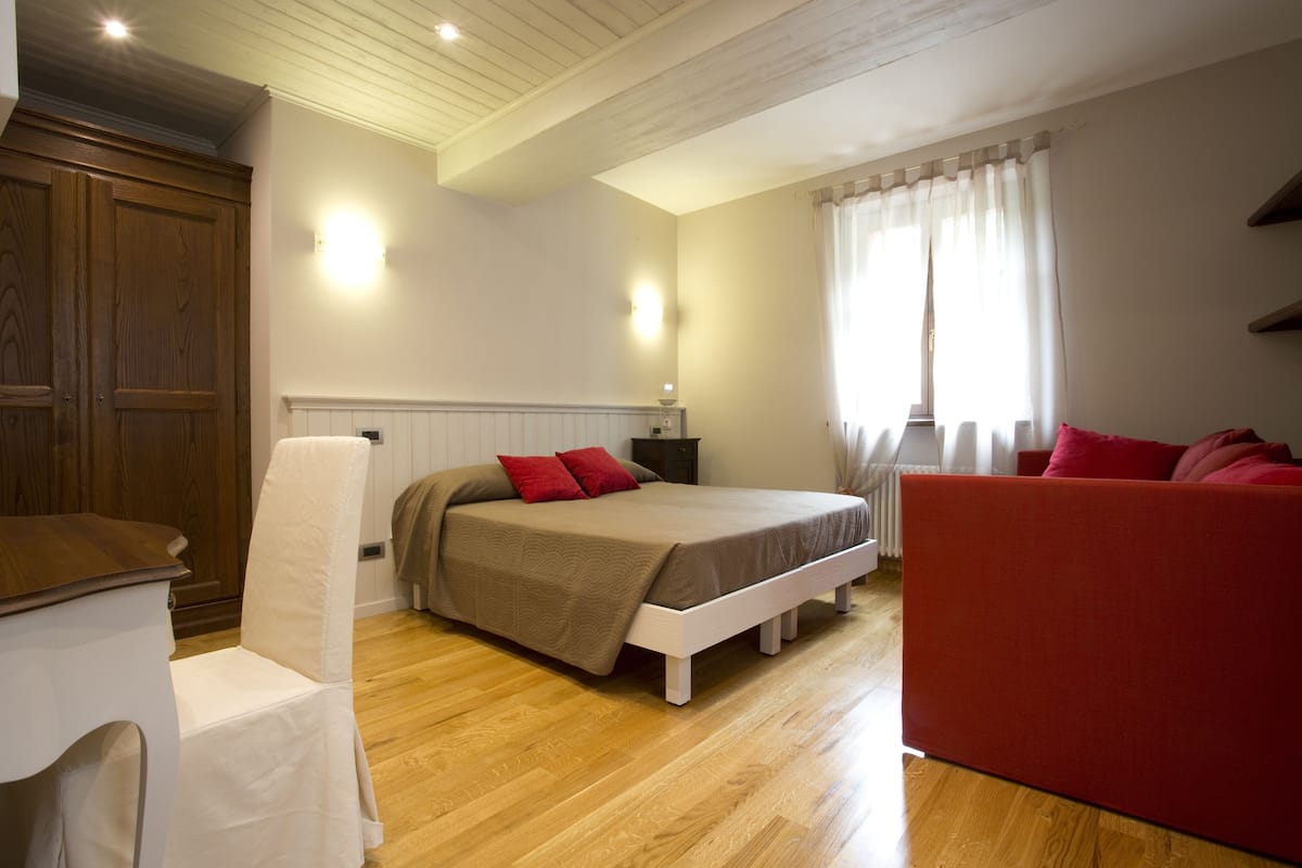 Ein Schlafzimmer mit einem Bett, einem Sessel, einem roten Sofa, Holzdielen und einem Kleiderschrank.