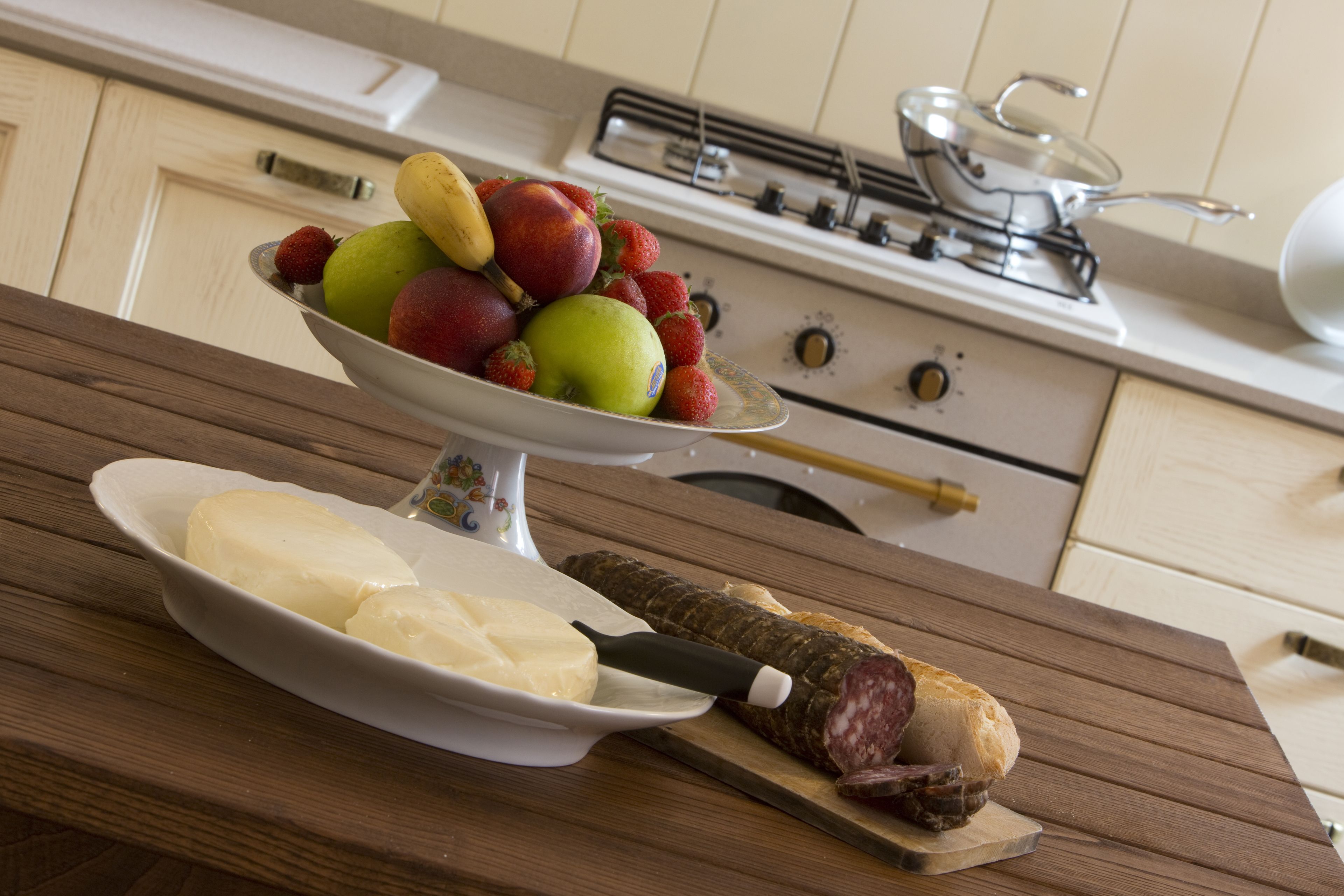 A kitchen counter with a fruit bowl, a plate of cheese, and a sliced salami on a cutting board.