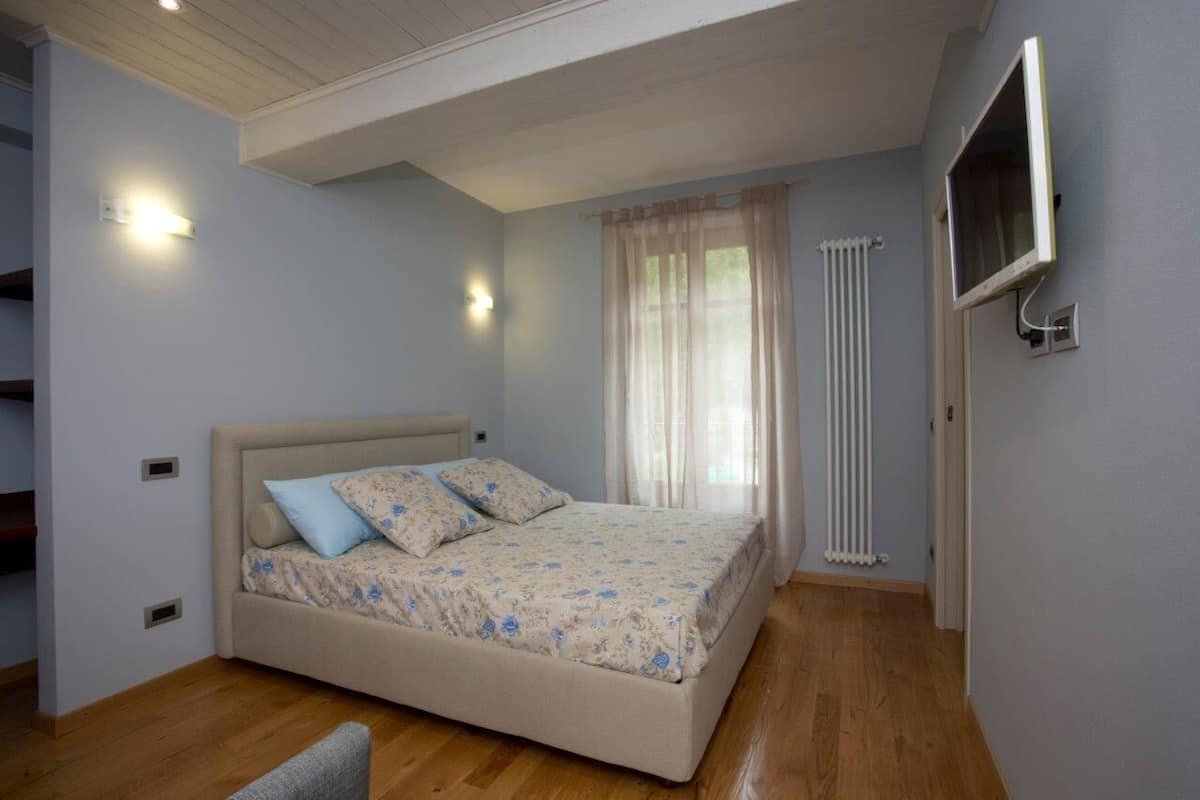 Ein Schlafzimmer mit Bett, einem an der Wand montierten Fernseher, einem Fenster mit Vorhängen und einer Heizung.