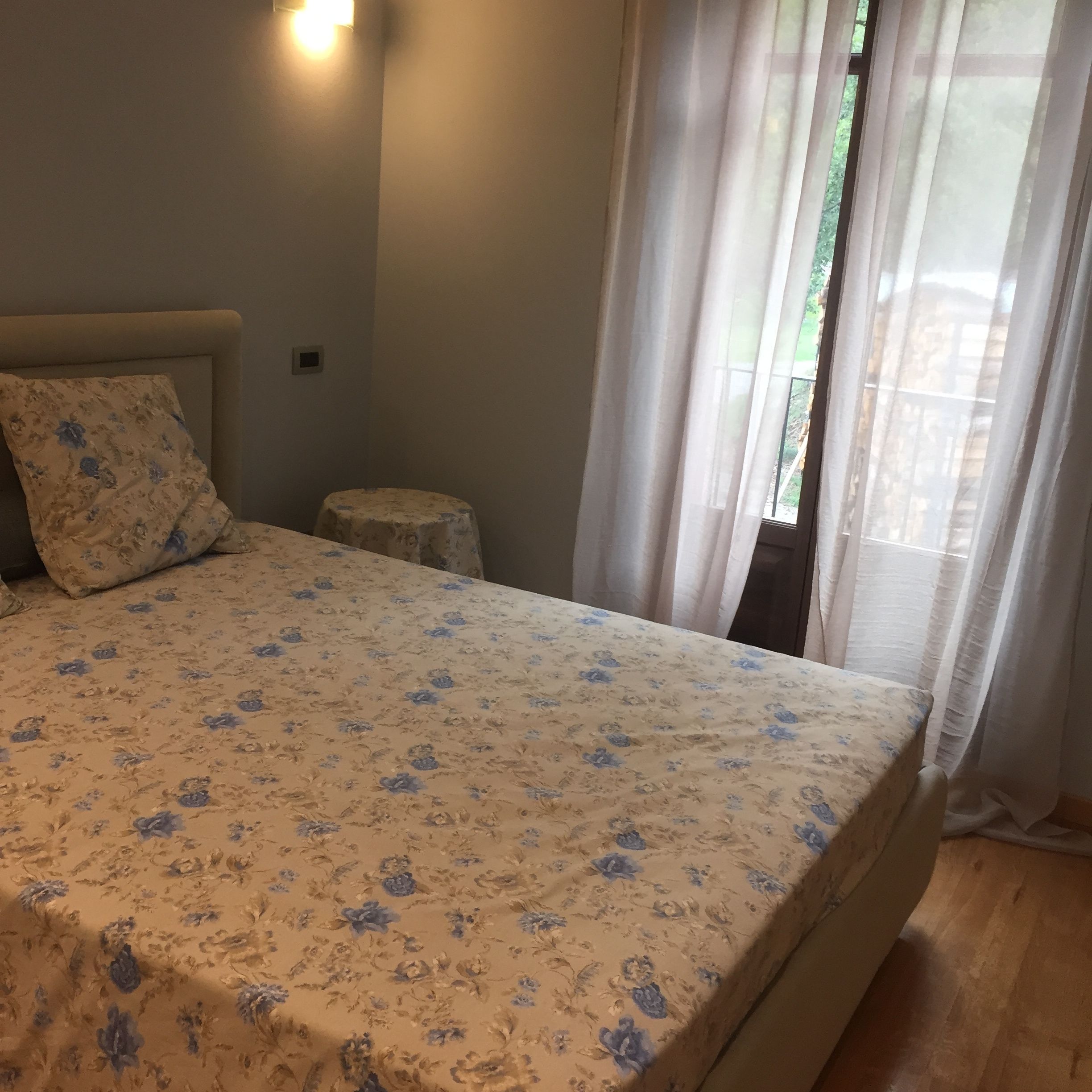 A bedroom with a bed, a nightstand, a wall-mounted light, and a window with curtains.