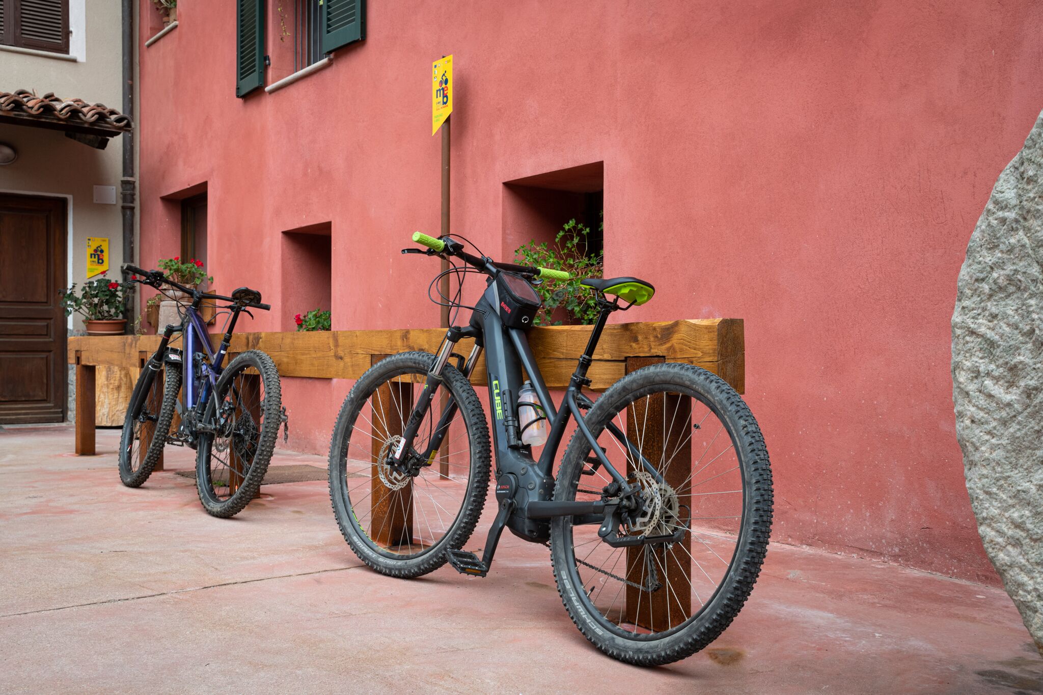 Two bicycles parked against a red wall with a wooden bench nearby.