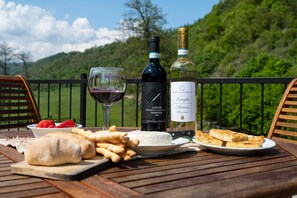 Una mesa con una botella de vino, una copa de vino tinto, una botella de vino blanco, un plato de queso, un tazón de fresas y pan, todo sobre una mesa de madera con una vista panorámica al fondo.