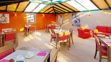Une salle à manger lumineuse, avec des tables en bois et des chaises rouges, agrémentée d’œuvres d’art aux murs et d’une verrière au plafond.