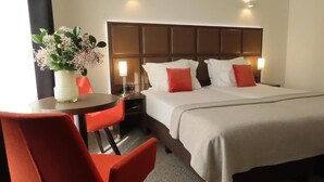 A hotel room with a bed, a nightstand, a table with a vase of flowers, and two red chairs. - Adagio hotel (Knokke-Heist)