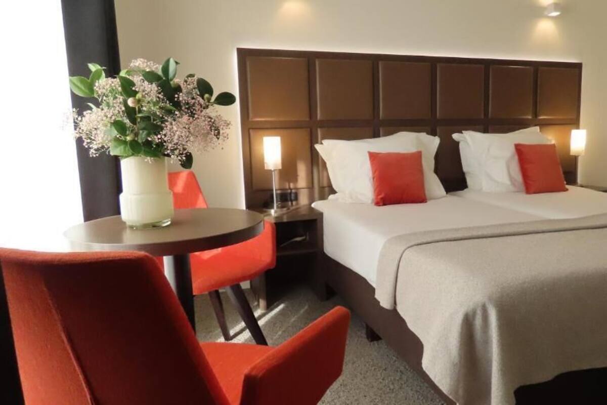 A hotel room with a bed, a nightstand, a table with a vase of flowers, and two red chairs.
