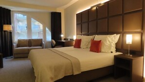 A hotel room with a large bed, a sofa, a desk, and a window with curtains. - Adagio hotel (Knokke-Heist)