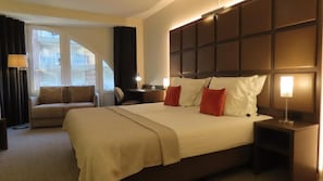 A hotel room with a large bed, a sofa, a desk, and a window with curtains. - Adagio hotel (Knokke-Heist)