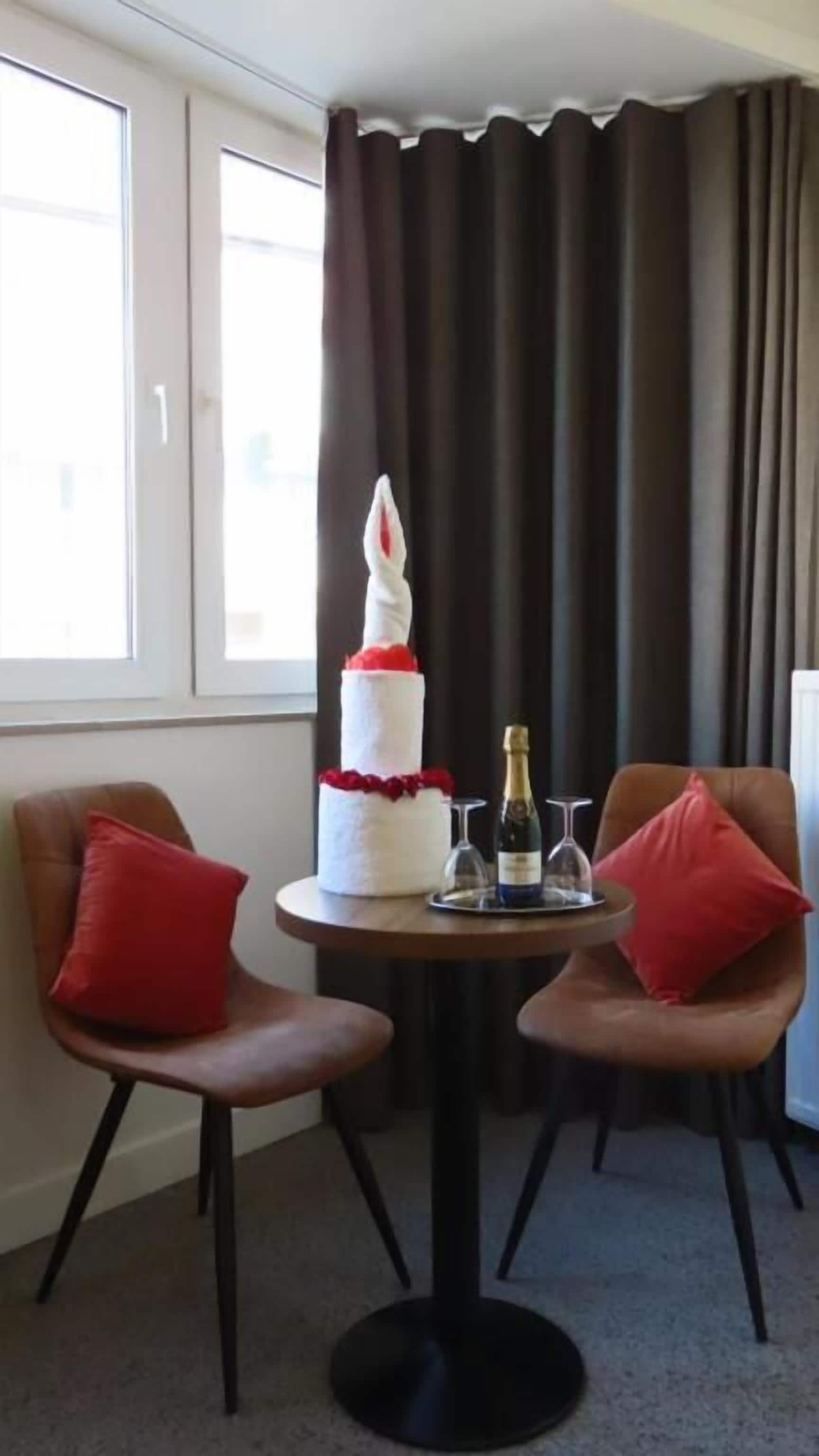 Una habitación con dos sillones de cuero marrón, una mesa redonda con un pastel blanco y champán, y vistas a un edificio a través de una ventana con cortinas marrones.