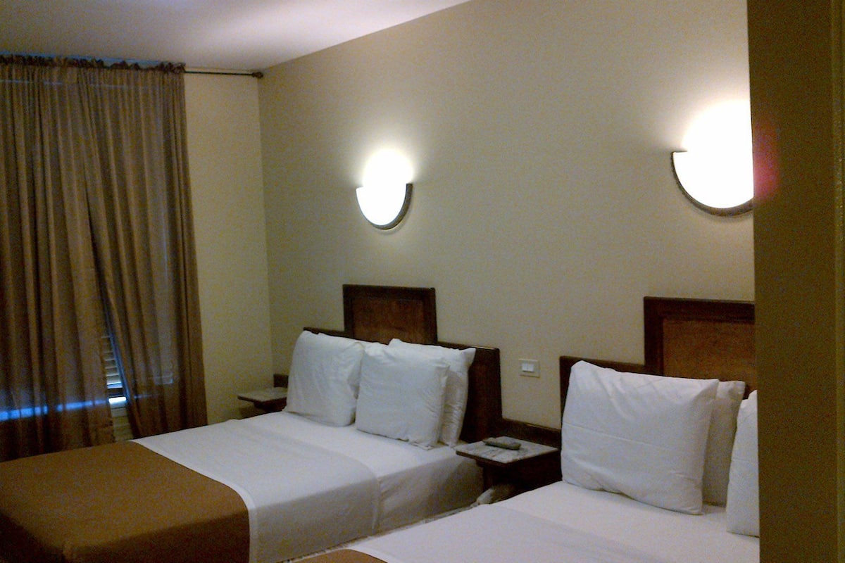 Una habitación de hotel con dos camas, una mesita de noche, una lámpara y un ventanal con cortinas.