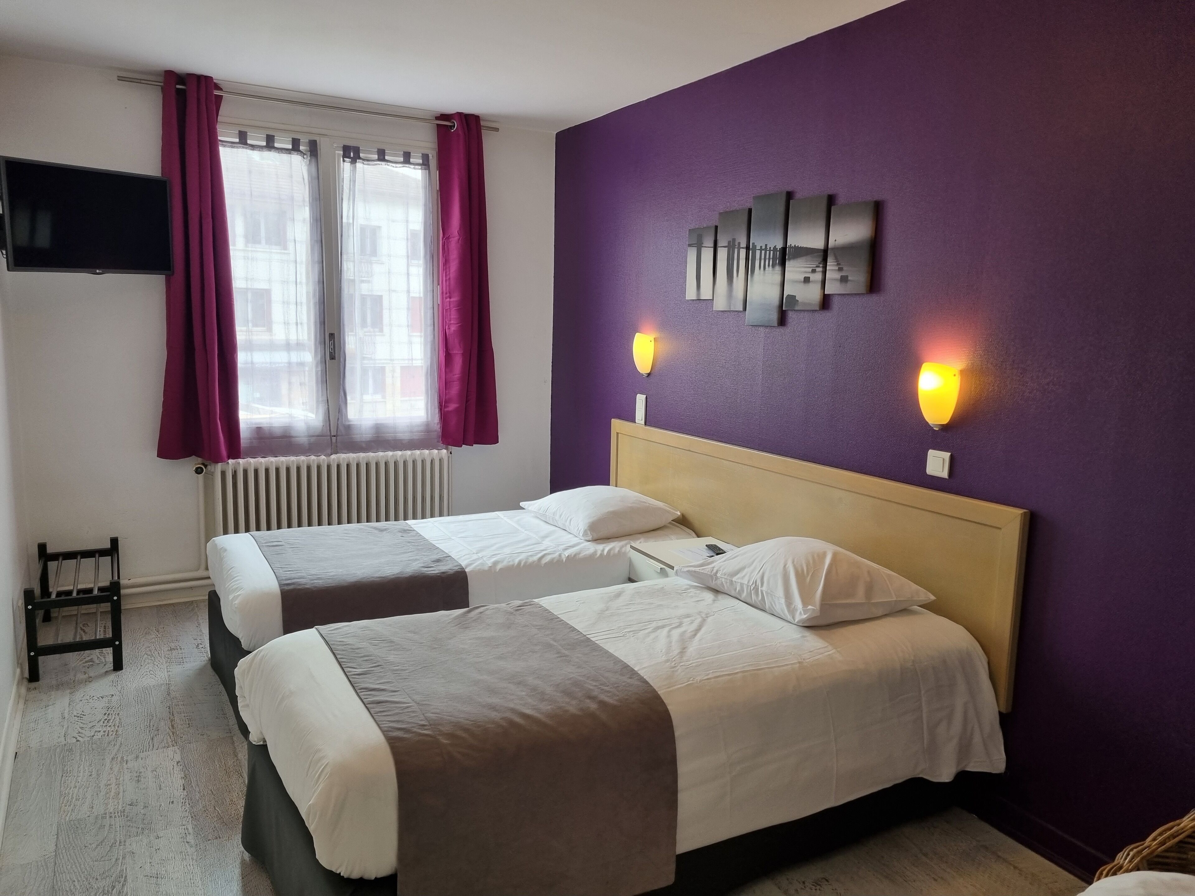 Habitación de hotel con dos camas, una ventana con cortinas, un televisor montado en la pared y dos apliques.