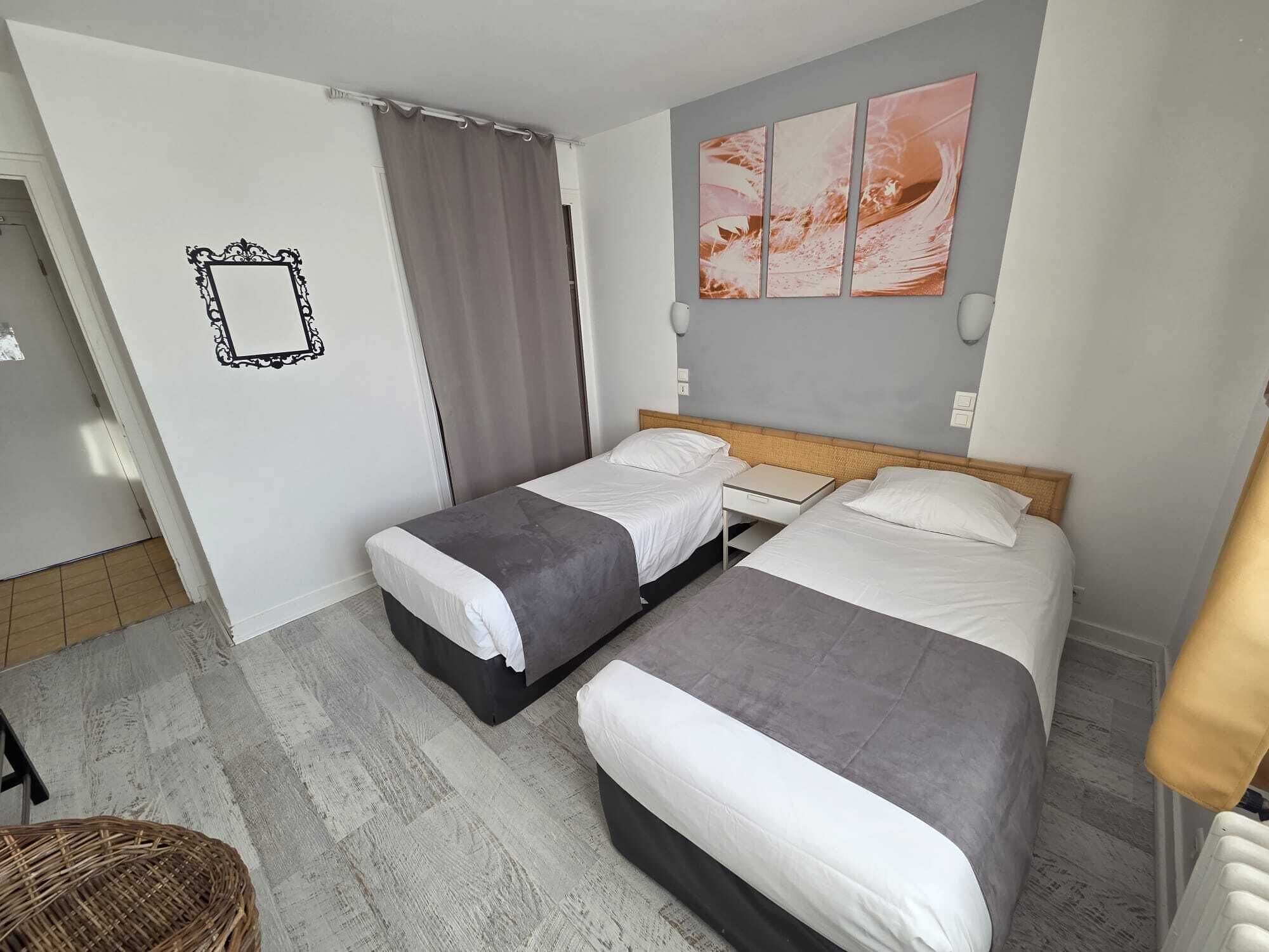 Habitación de hotel con dos camas, una cortina gris, un cuadro colgado en la pared y una silla de mimbre.
