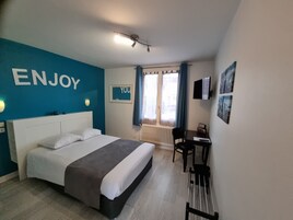 Quarto de hotel com cama, escrivaninha, cadeira, radiador e janela com cortinas.