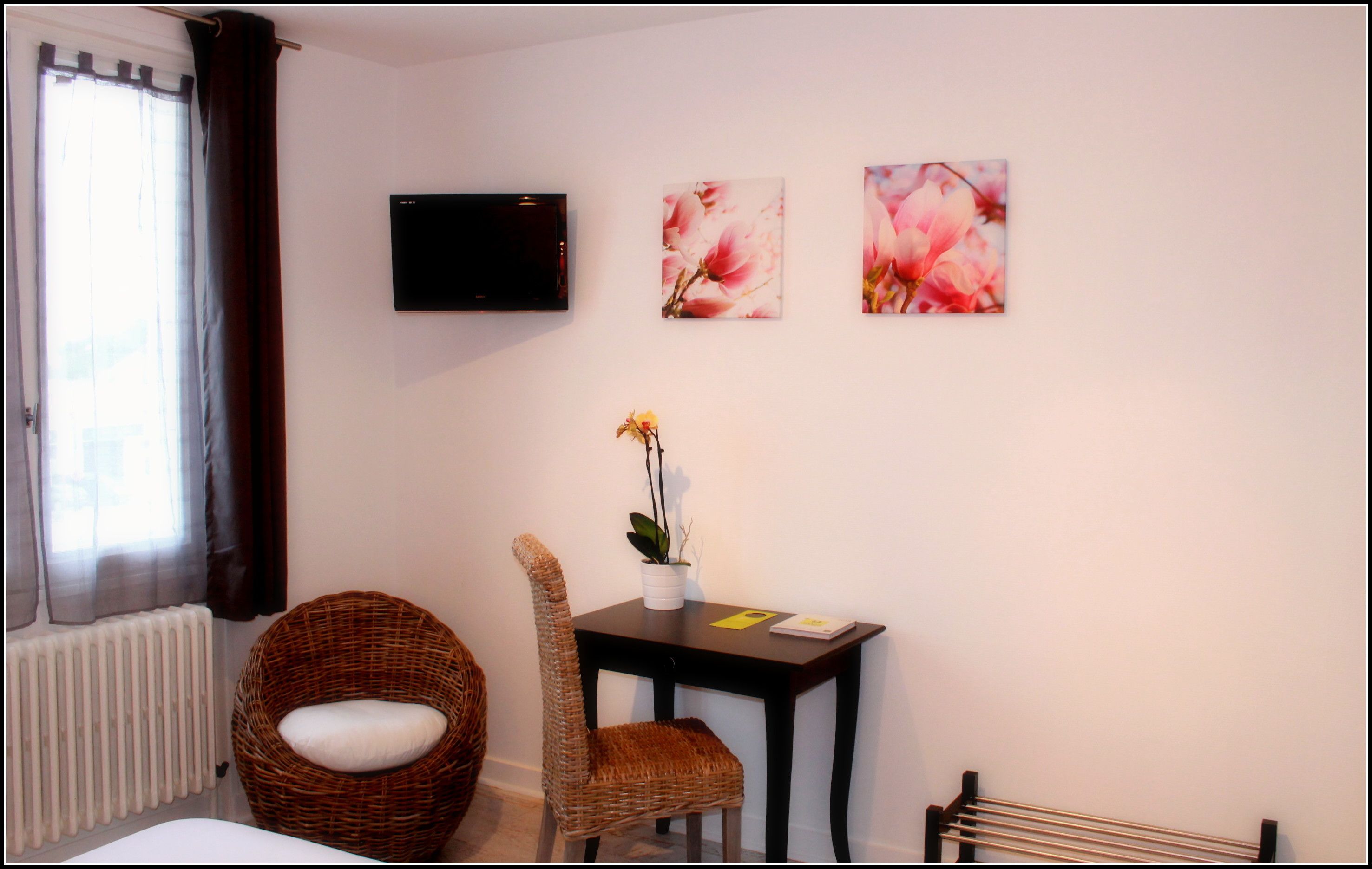 A hotel room with a wicker chair, a small table with a vase of flowers, and two wall-mounted pictures.