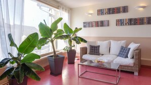 Een woonkamer met een witte bank, twee potplanten, een glazen salontafel en een boekenkast vol boeken.
