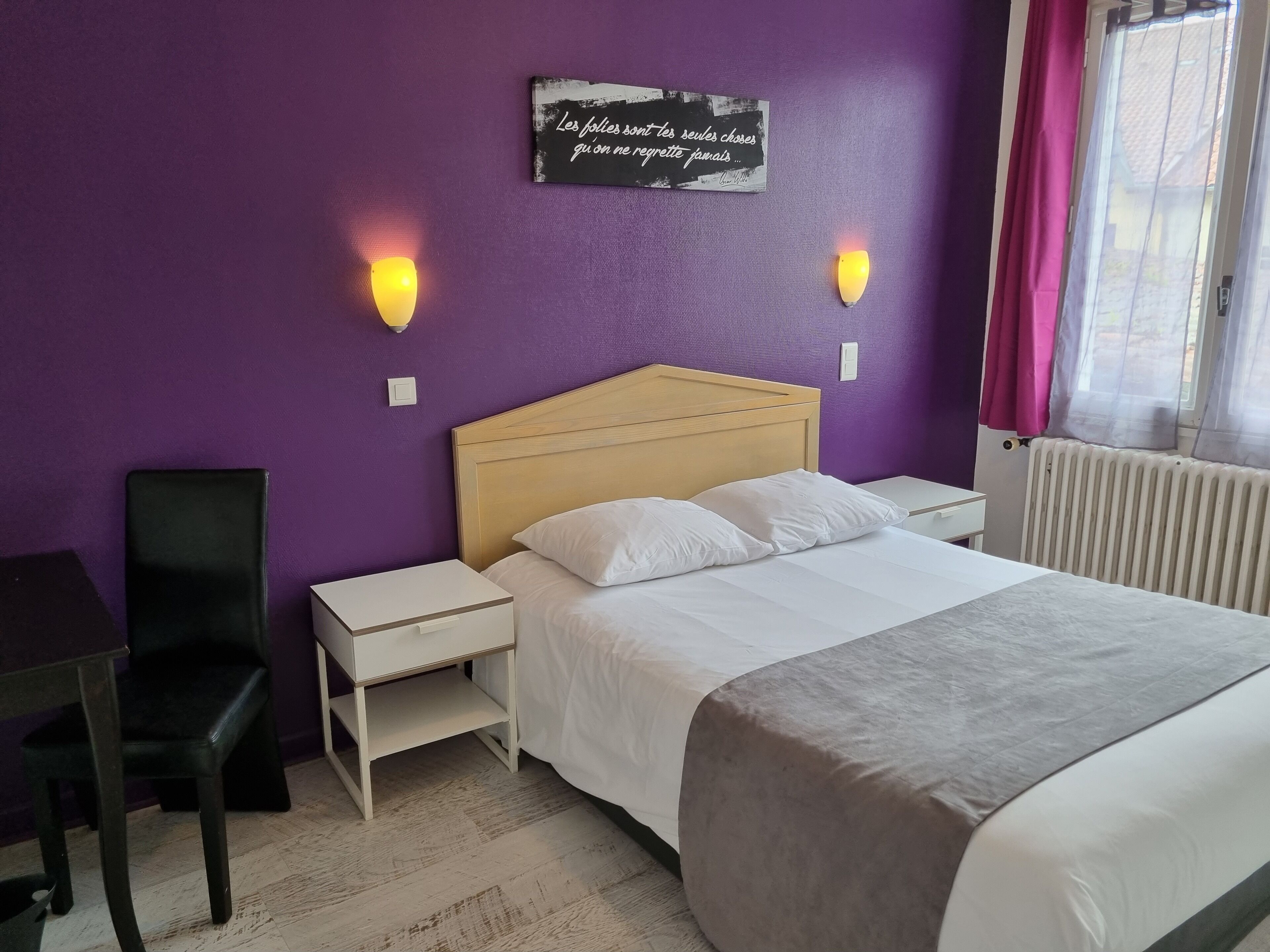 Una habitación de hotel con una pared morada, cabecera de madera, cama blanca, mesita de noche, escritorio, silla y ventana con cortinas.