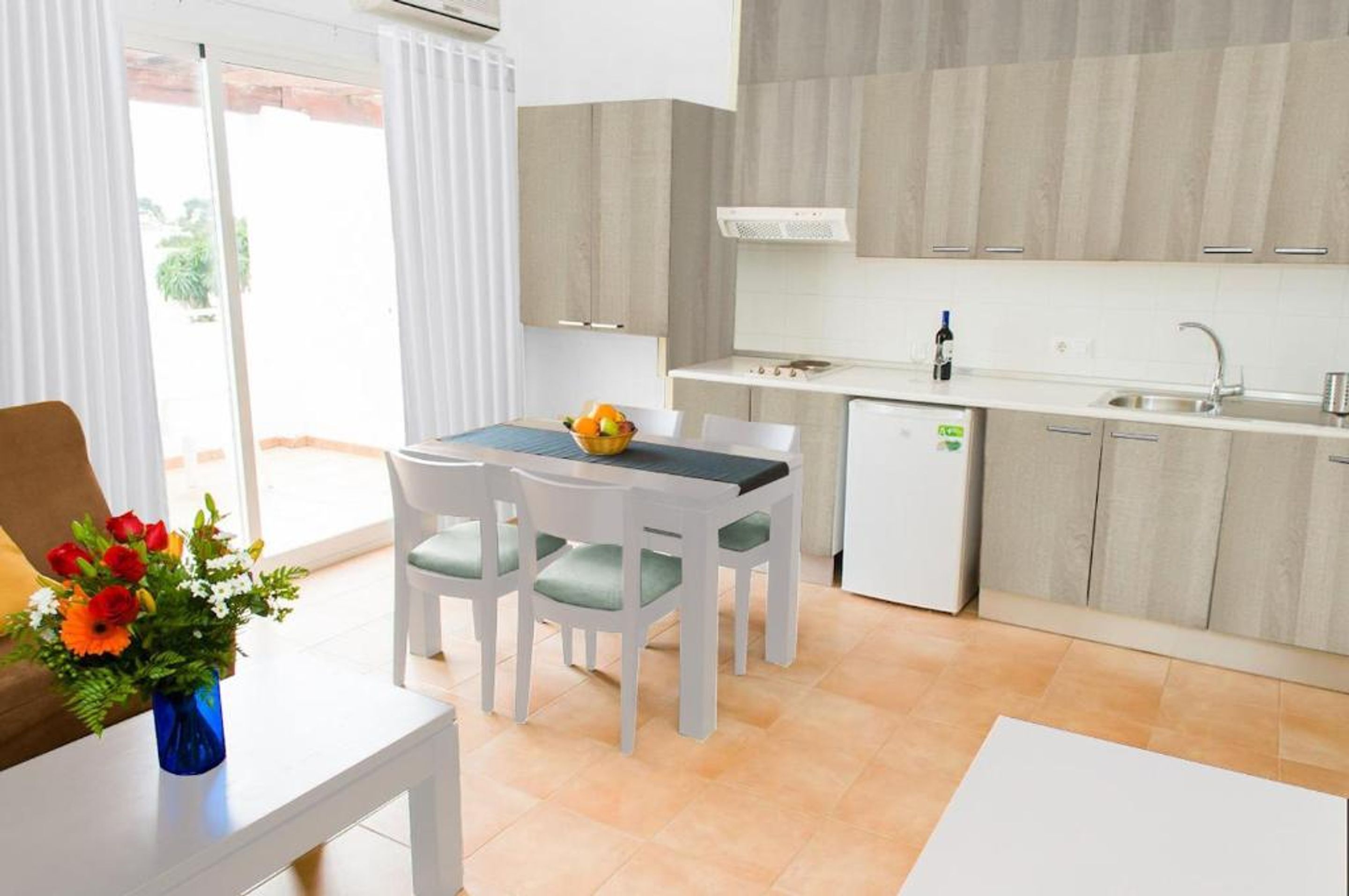 A modern kitchen with white cabinets, a built-in oven, and a sink. There is a dining table with chairs and a vase of flowers.