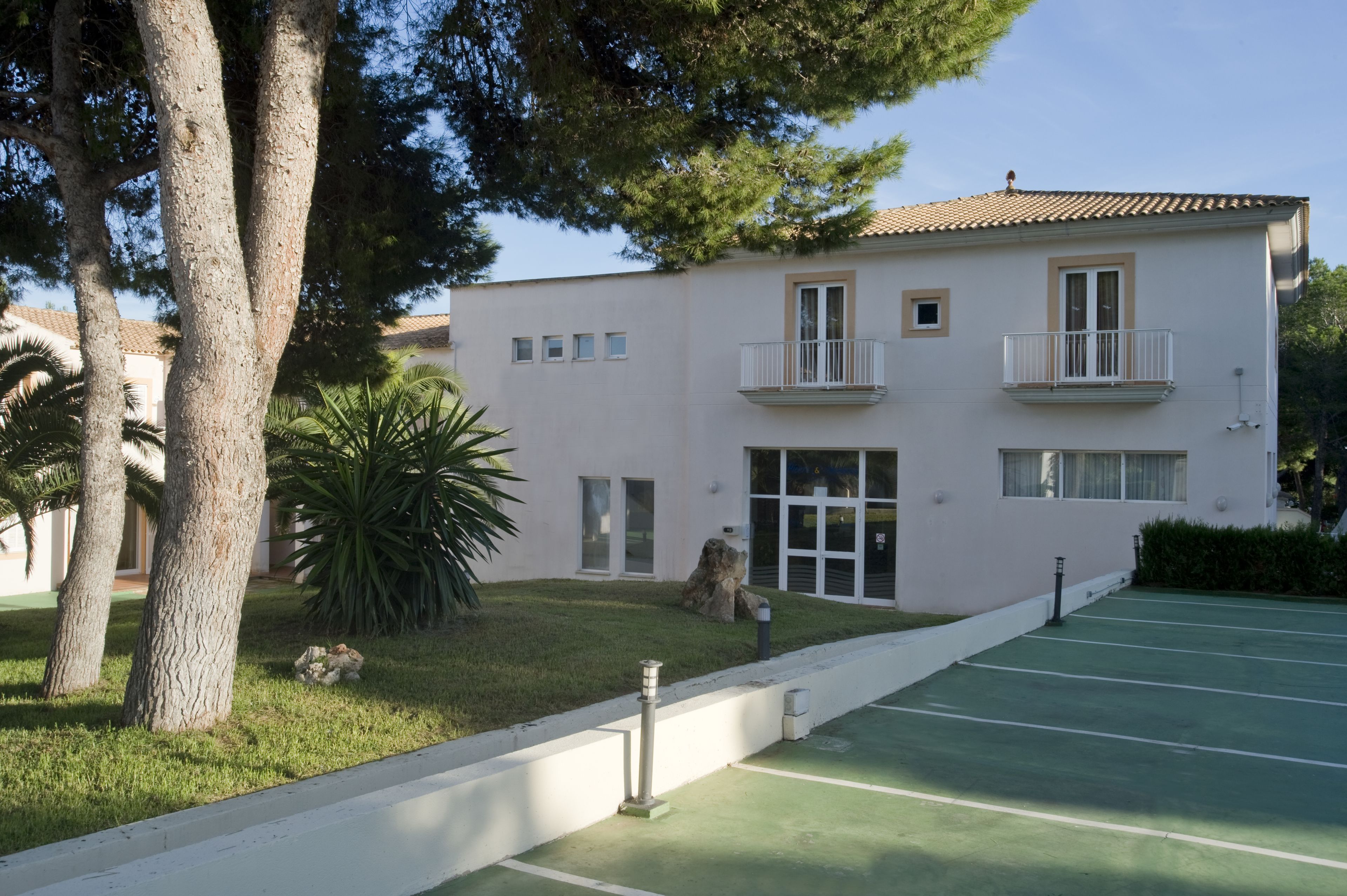 Un edificio de dos plantas con balcón, un jardín pequeño y una cancha de tenis.