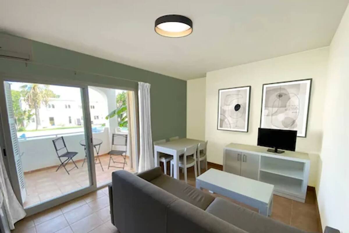 A living room with a sofa, a TV, a dining table with chairs, and a balcony with a view of buildings and greenery.