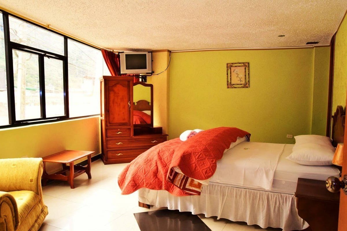 Une chambre à coucher avec un lit, une armoire en bois, un téléviseur posé sur un meuble, une petite table et une fenêtre donnant sur l’extérieur.