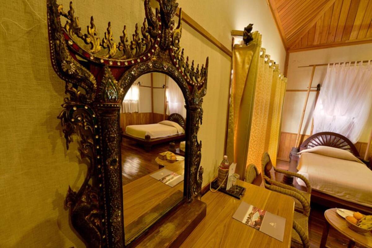 A traditional room with a large ornate mirror, a bed with a wooden headboard, a small table with a book, and a chair.