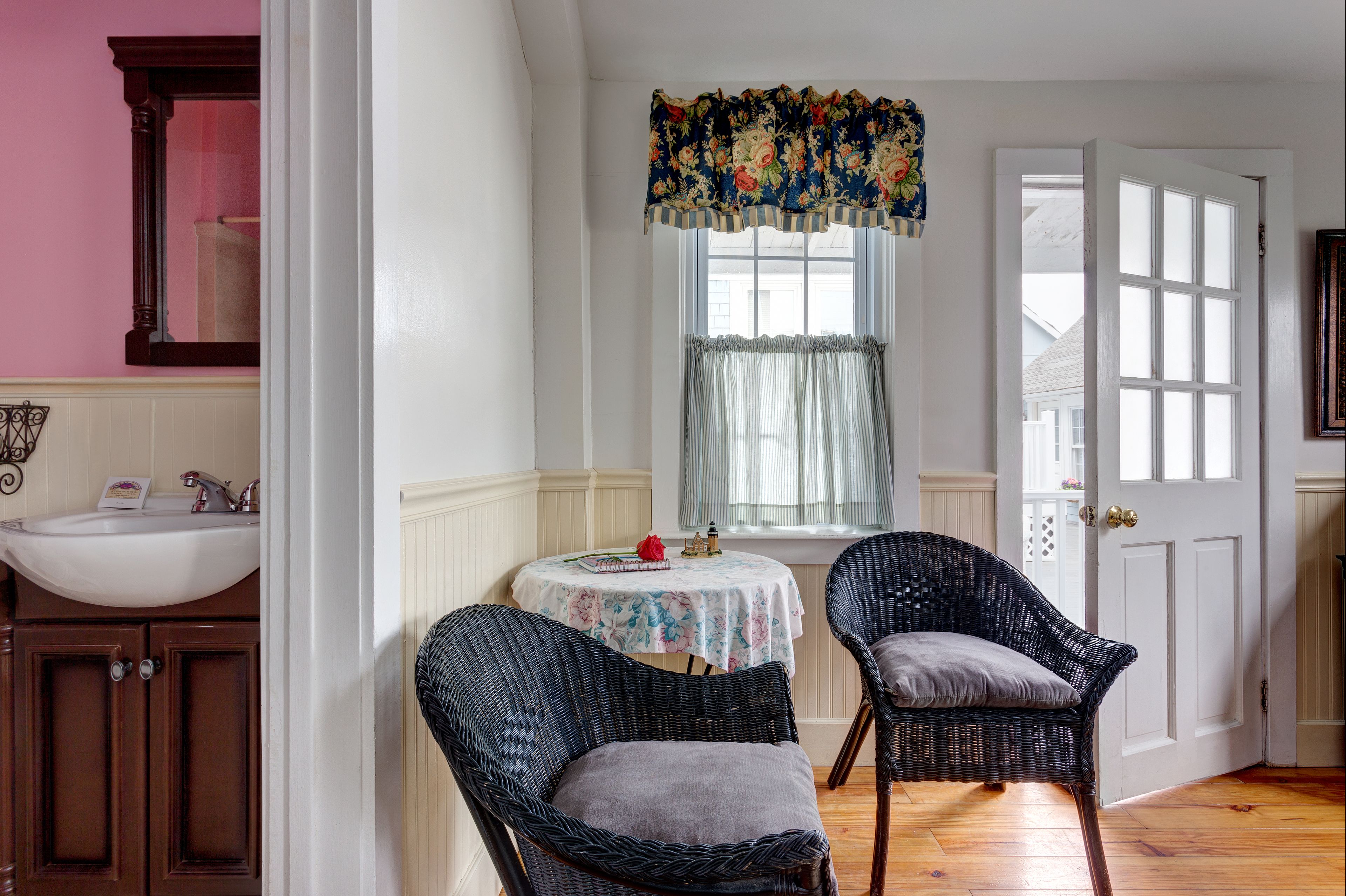 A room with a white sink, wooden cabinets, two wicker chairs, a small table with a floral tablecloth, and a window with a floral valance.