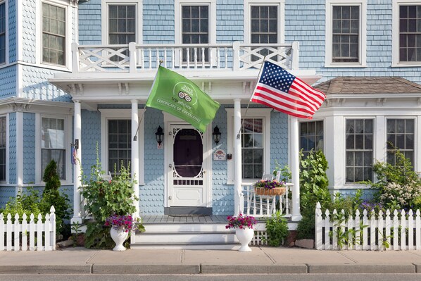 Una casa azul con un porche blanco, una bandera verde y una bandera estadounidense.
