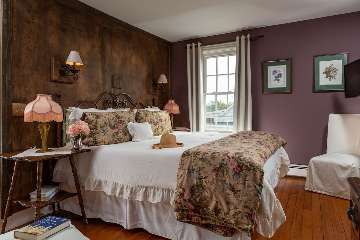 Une chambre à coucher avec une tête de lit en bois, un lit recouvert d’une literie à motifs floraux, une table de chevet avec une lampe et une chaise.