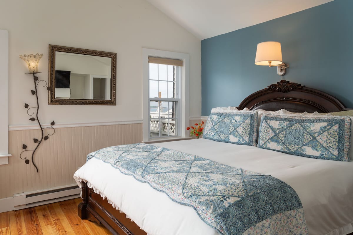 Une chambre avec un grand lit, une table de chevet, une lampe fixée au mur, un miroir et une vue sur l’extérieur par une fenêtre.
