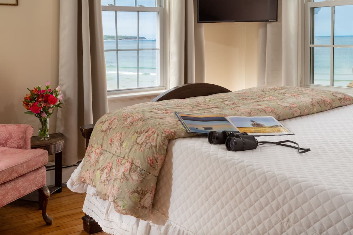 Une chambre à coucher avec un lit, une chaise, une table ornée d’un vase de fleurs et une fenêtre donnant sur la mer.