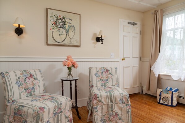 A room with two upholstered chairs, a small table with a vase of flowers, a wall-mounted lamp, and a door with a window. - The Blue Dory Inn (Block Island)