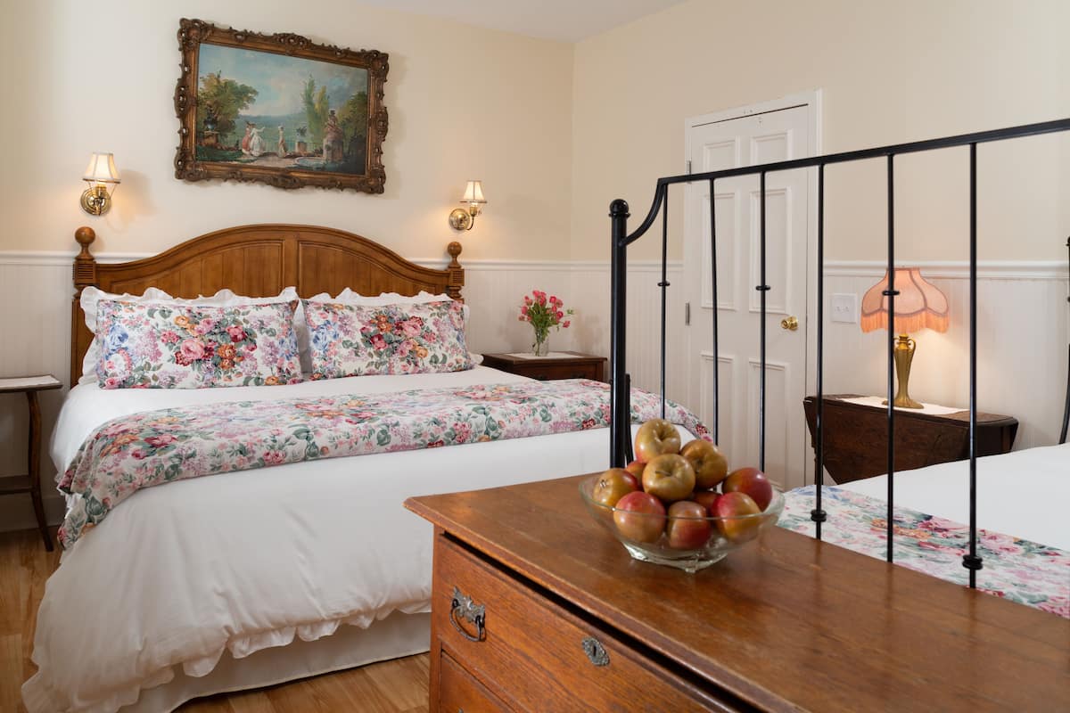 Une chambre à coucher avec un lit en bois, une literie à motifs floraux, une table de chevet avec une lampe, une commode sur laquelle est posé un panier de fruits, et une porte avec un cadre blanc.