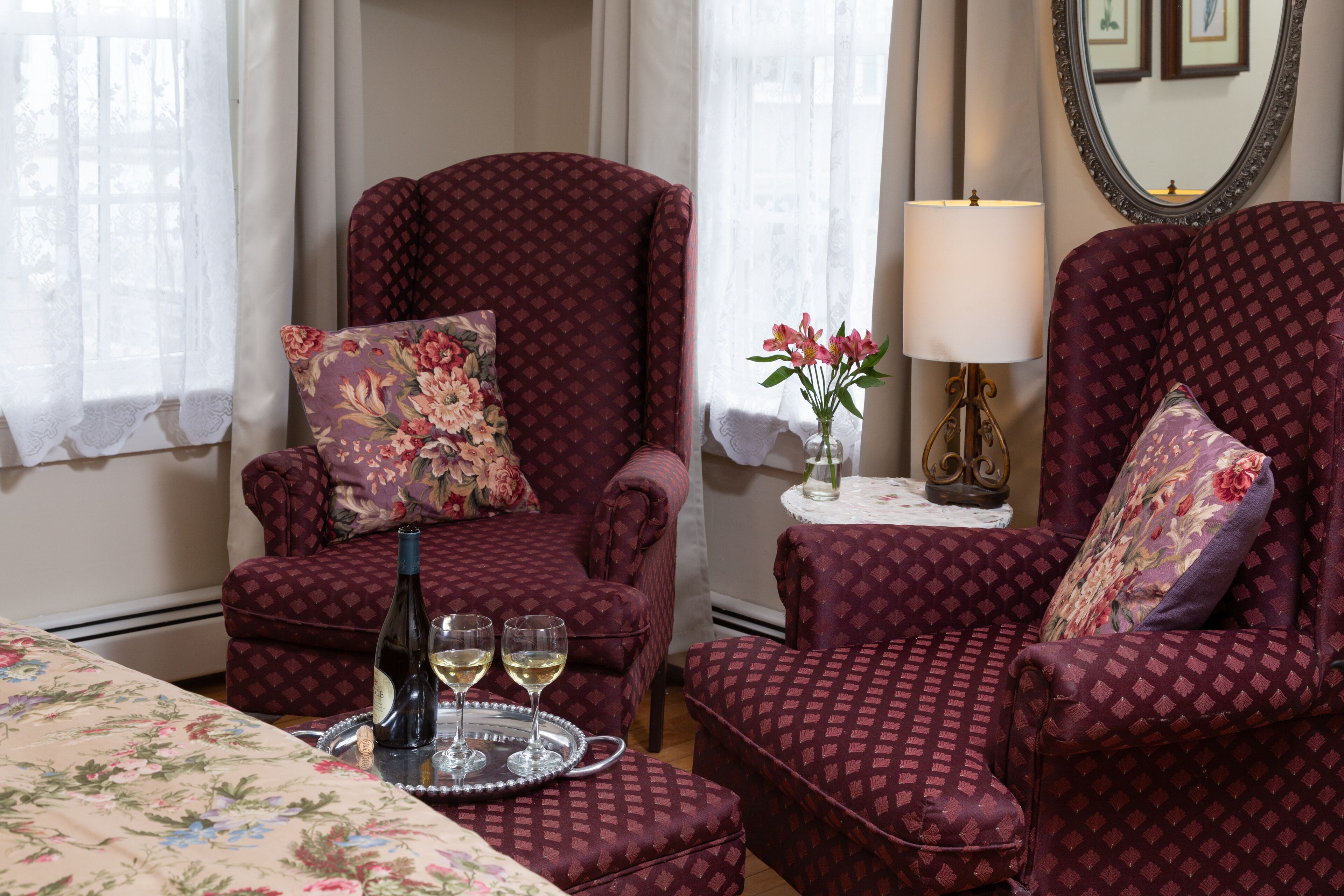 A room with two patterned armchairs, a floral bedspread, a table with a bottle and glasses, and a vase with flowers.