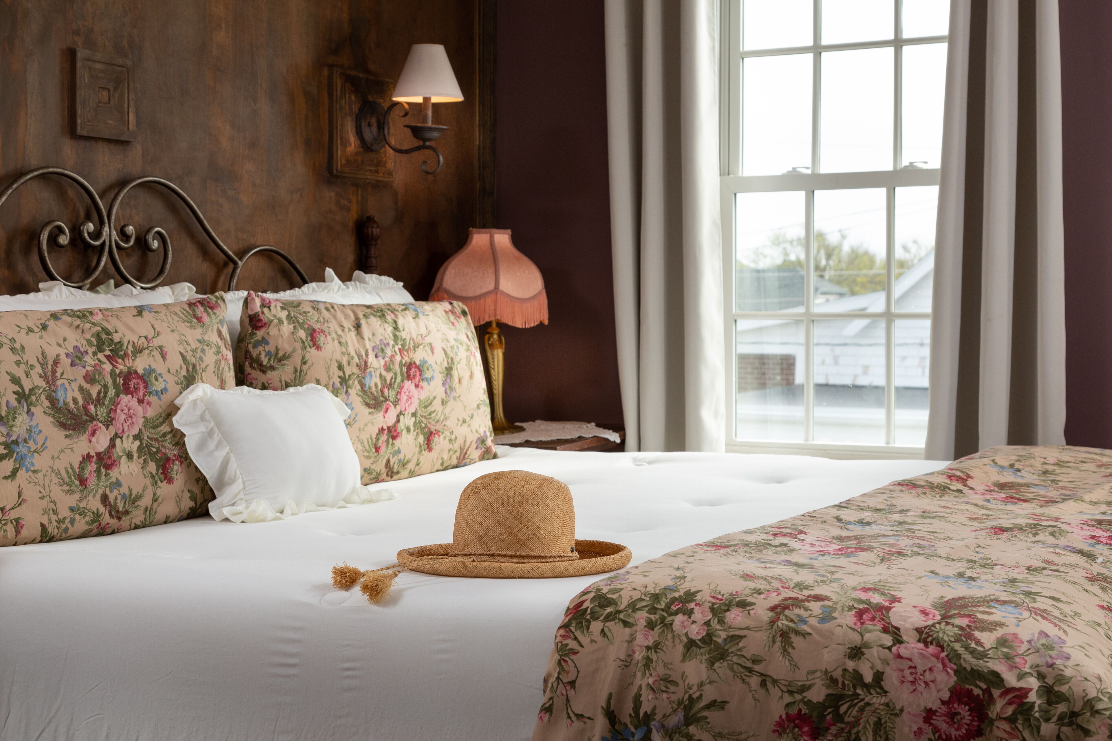 Un dormitorio con ropa de cama floral, una cama con cabecera, una lámpara de noche y una ventana con cortinas.