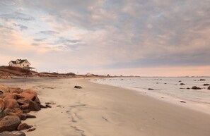 A beach with a house on a hill, rocky shoreline, and a cloudy sky.