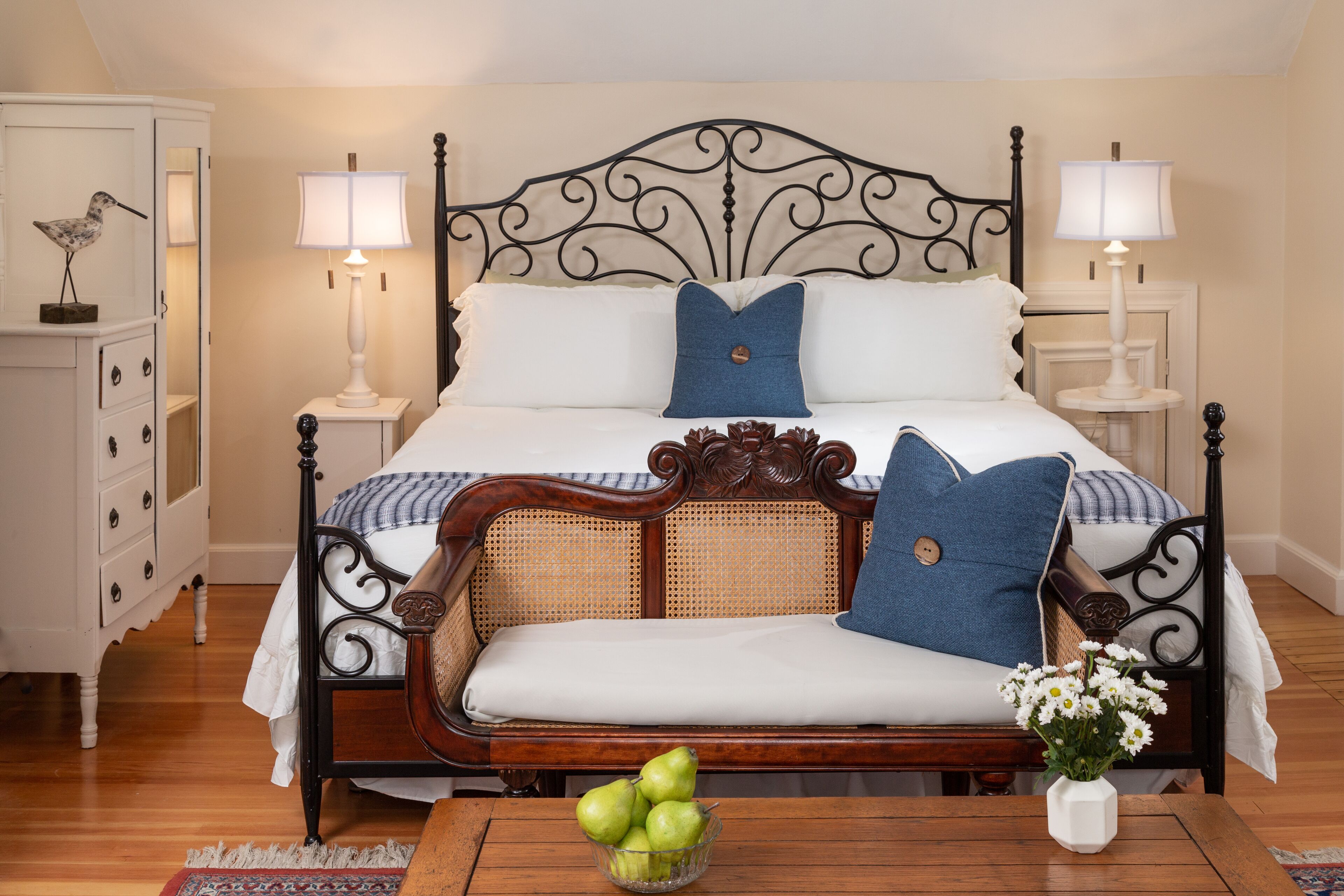A neatly arranged bedroom with a four-poster bed, bedside tables with lamps, a wooden dresser, and a small table with a fruit bowl and flowers.