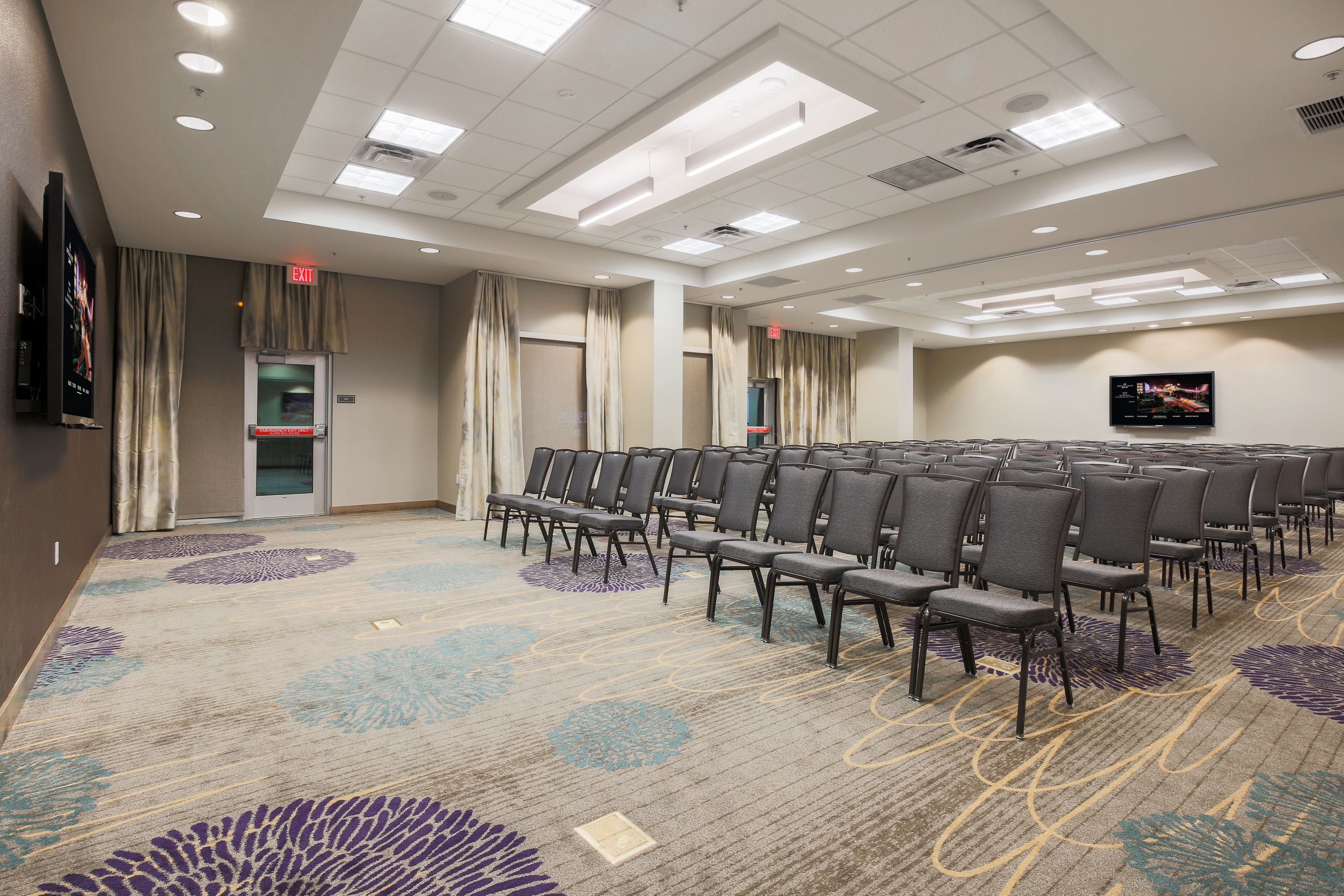 A conference room with rows of chairs, a carpeted floor, and a flat-screen TV on the wall.