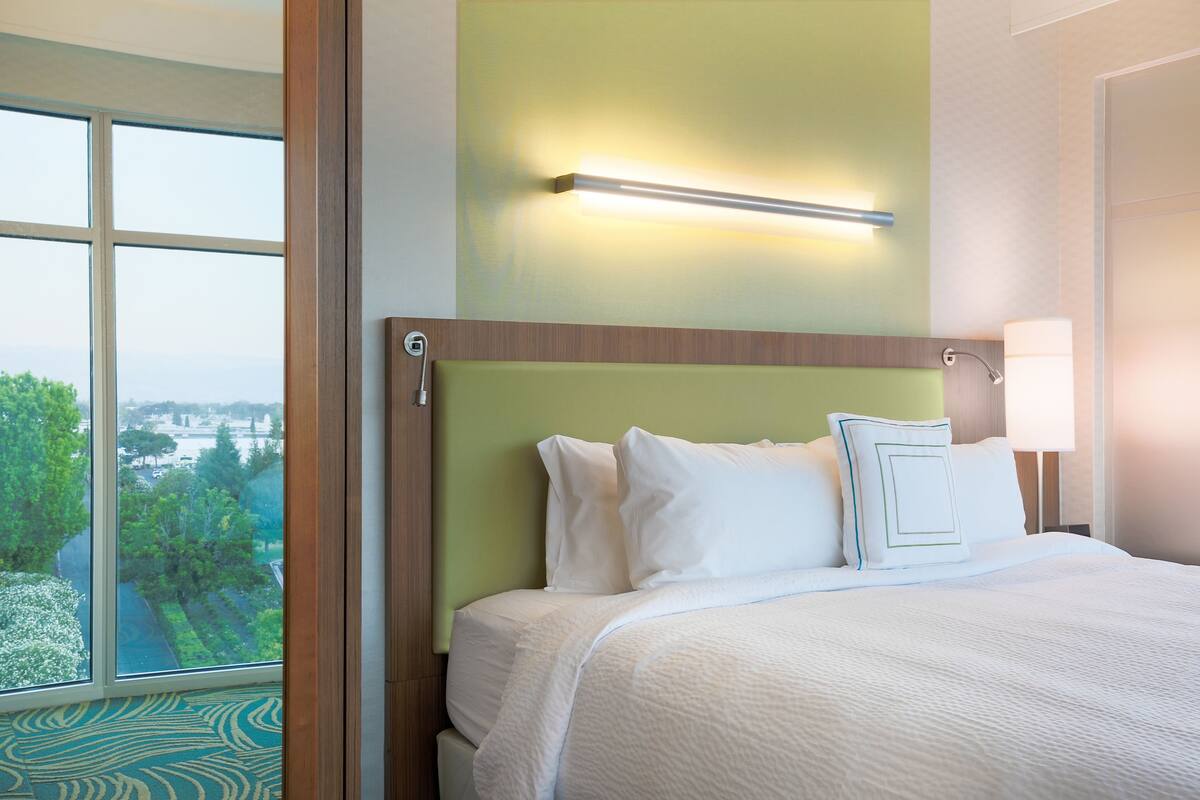 A hotel room with a large window, a bed with white bedding, a green accent wall, and a bedside lamp.